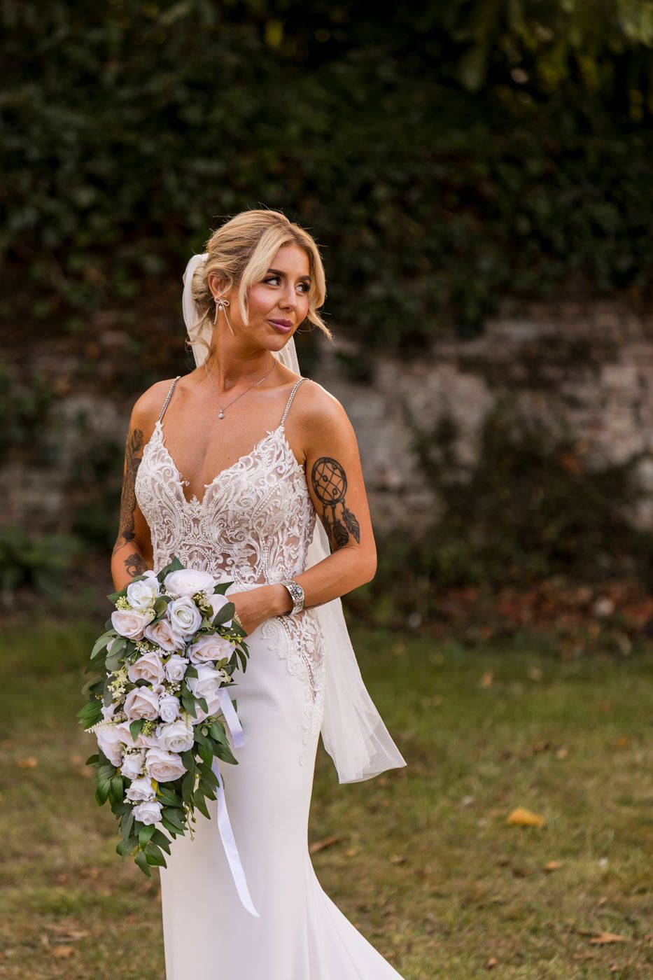 A bride in a fitted lace wedding dress holds a cascading bouquet of white and purple flowers in an outdoor garden setting.