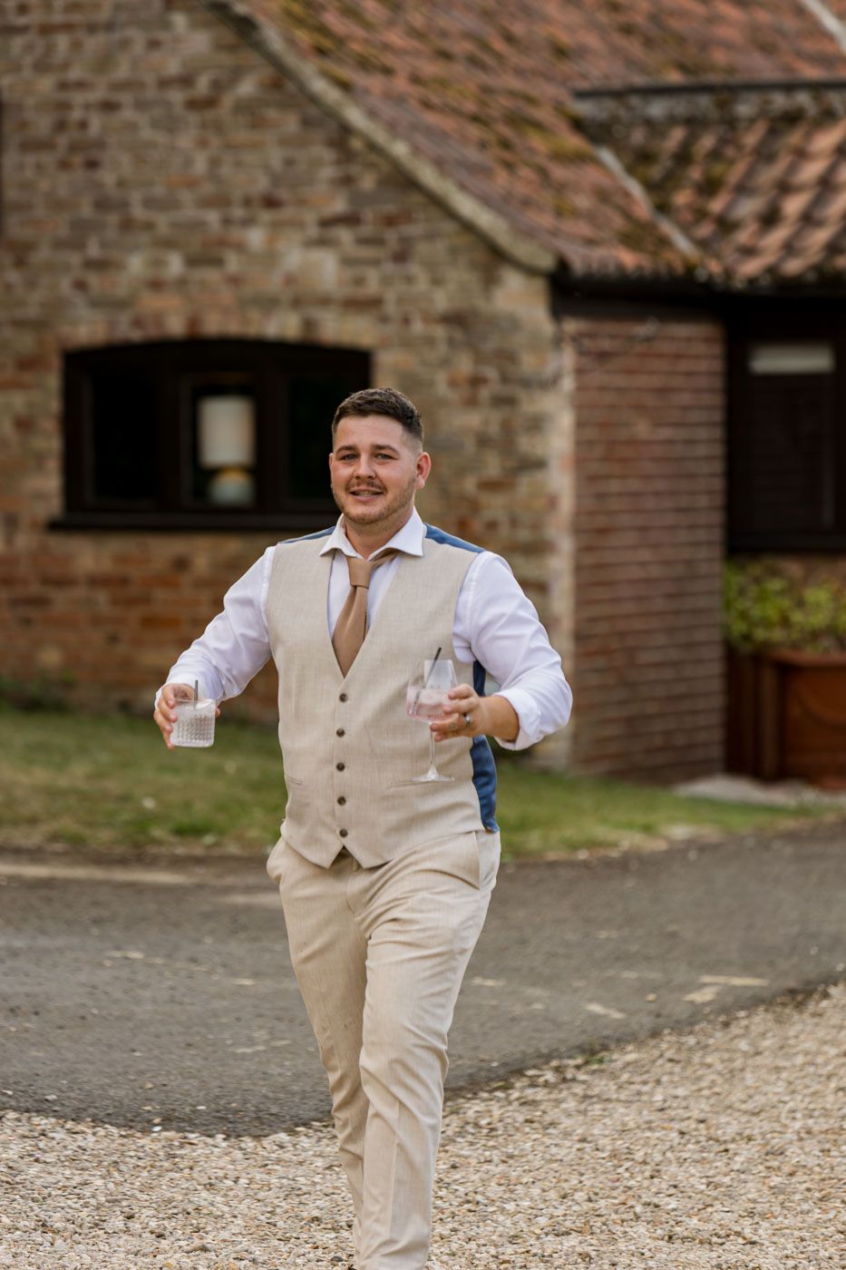 A groom in beige vest and pants runs joyfully across a gravel path near a brick building.