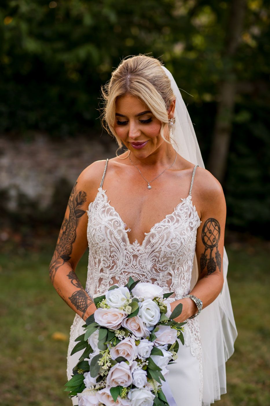 A bride with tattoos wears a lace wedding gown and holds a white and purple cascading bouquet.