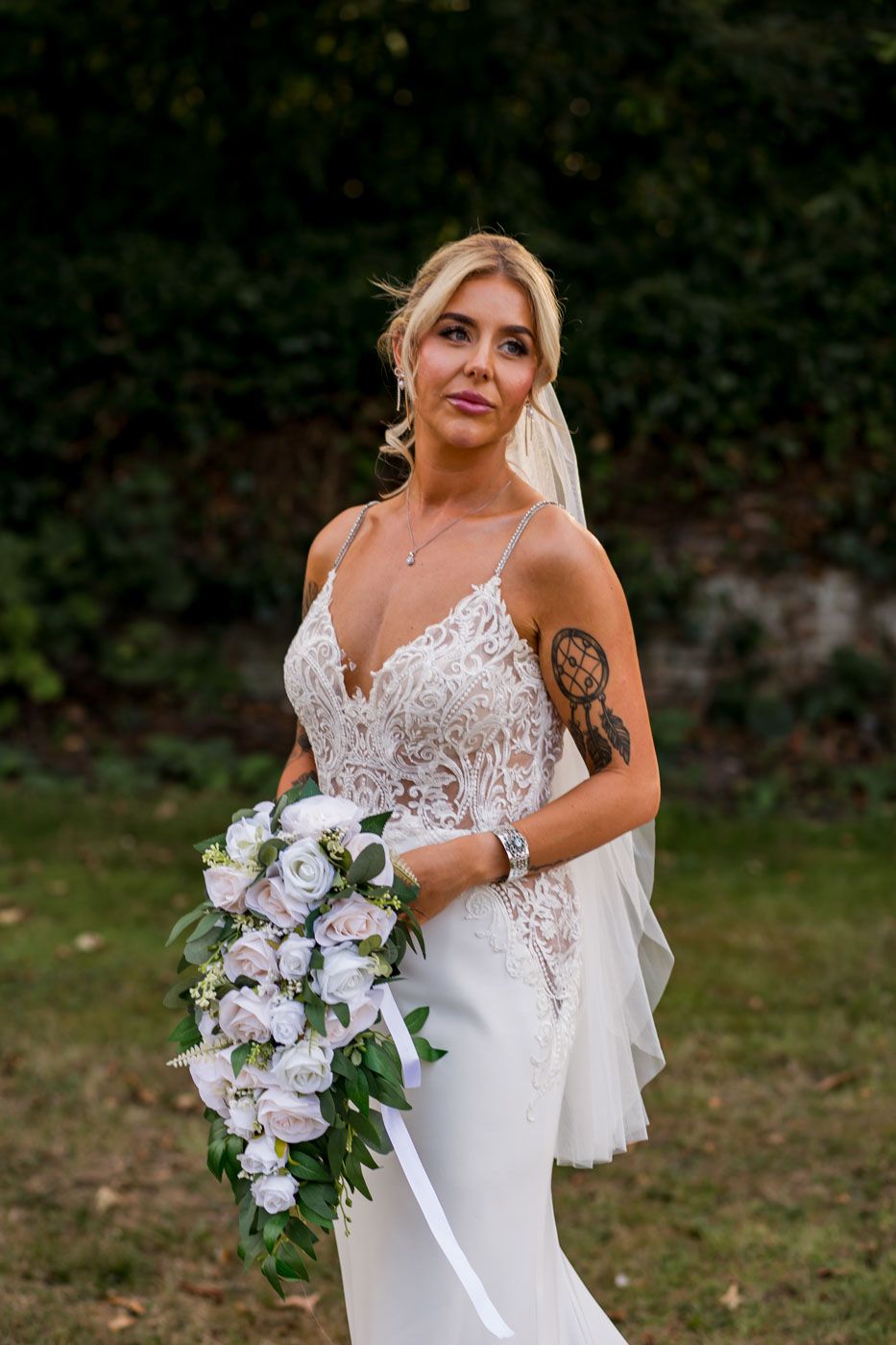 A bride in a form-fitting lace gown poses with an elegant cascading bouquet against a garden backdrop.