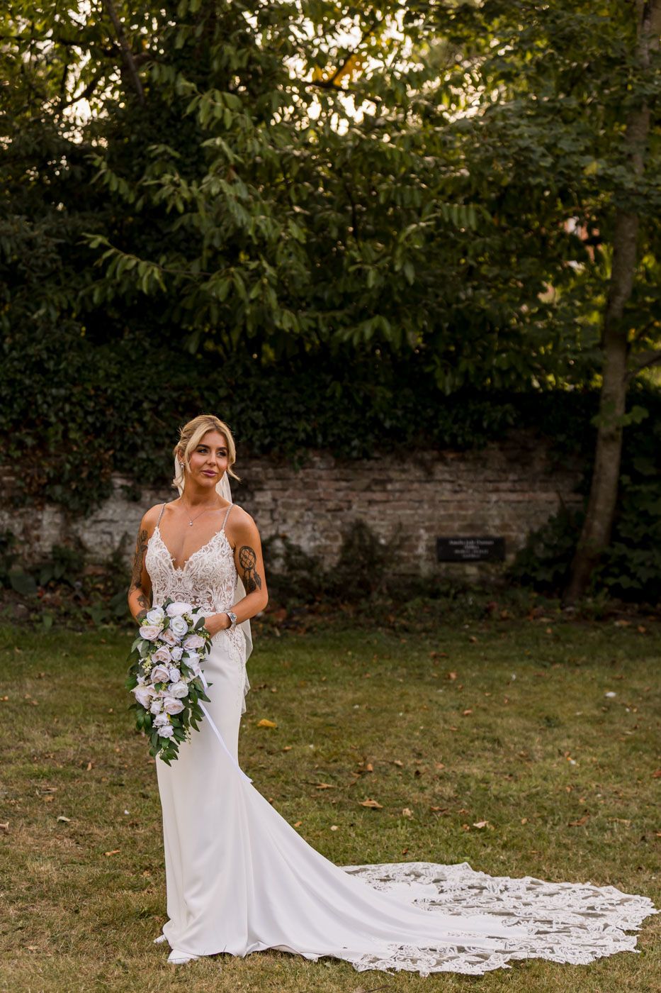 A bride in a sleek white gown with long train poses in a garden setting at sunset.