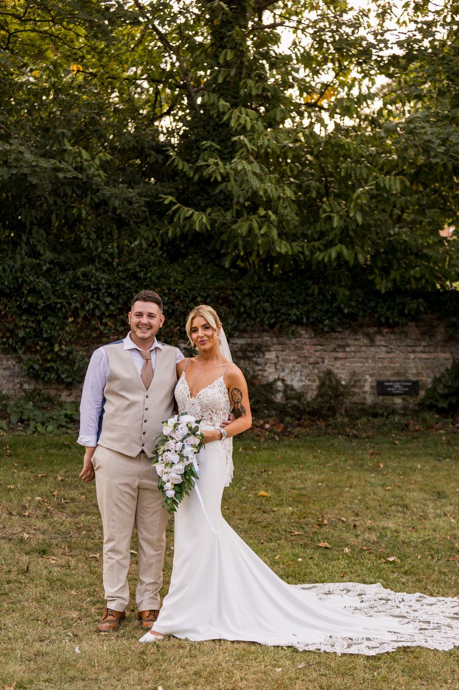 A wedding couple poses together on a lawn with brick wall and trees in the background.