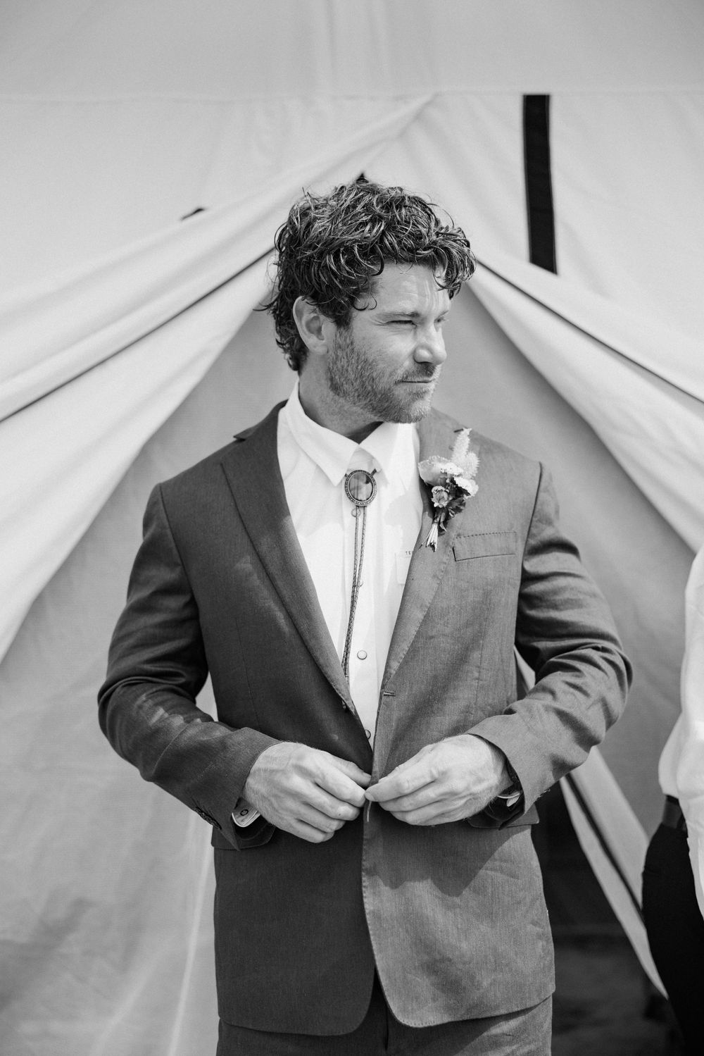 A groom in a gray suit and bolo tie stands in front of a white tent backdrop at a rustic wedding.