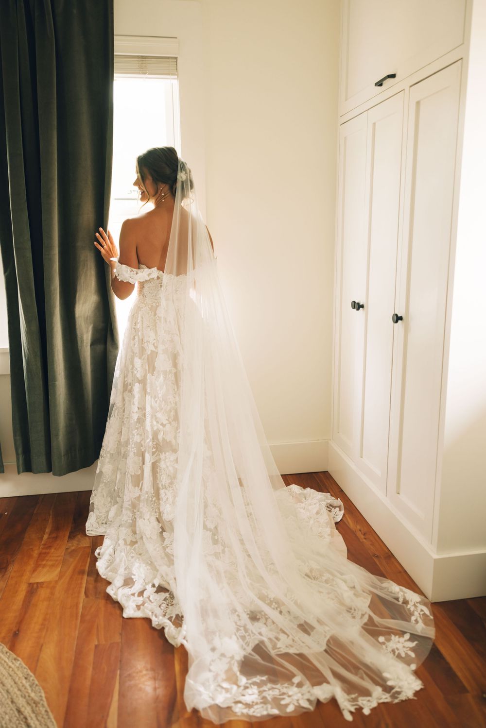 A flowing white lace wedding dress with a long train displayed on hardwood floors.