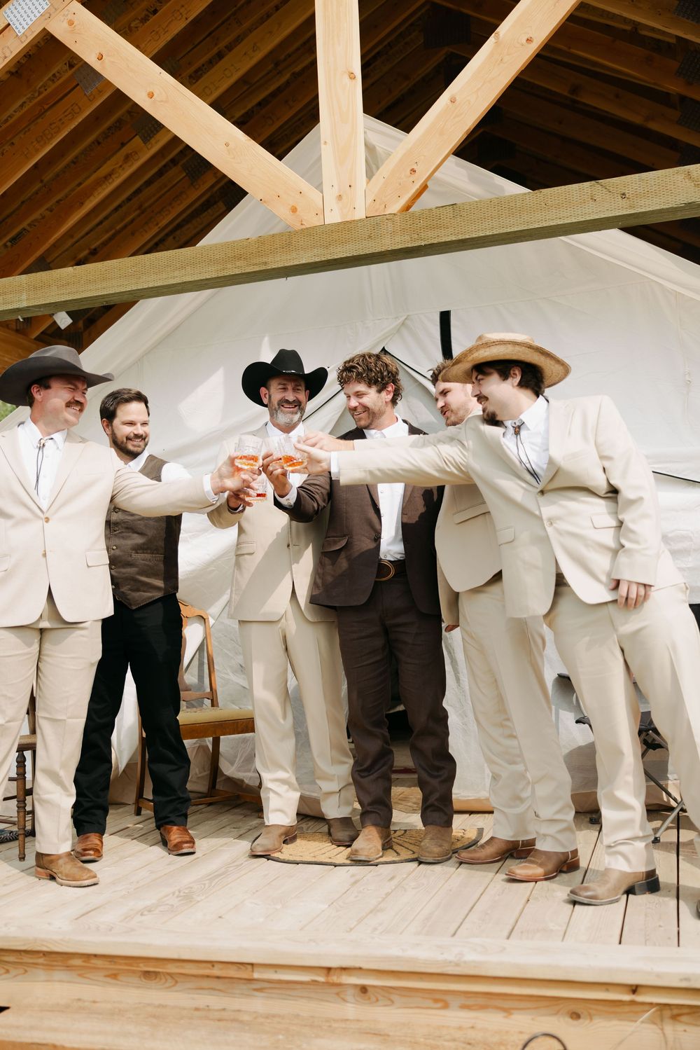 Wedding party in light colored attire lined up along wooden beams under a rustic barn ceiling.