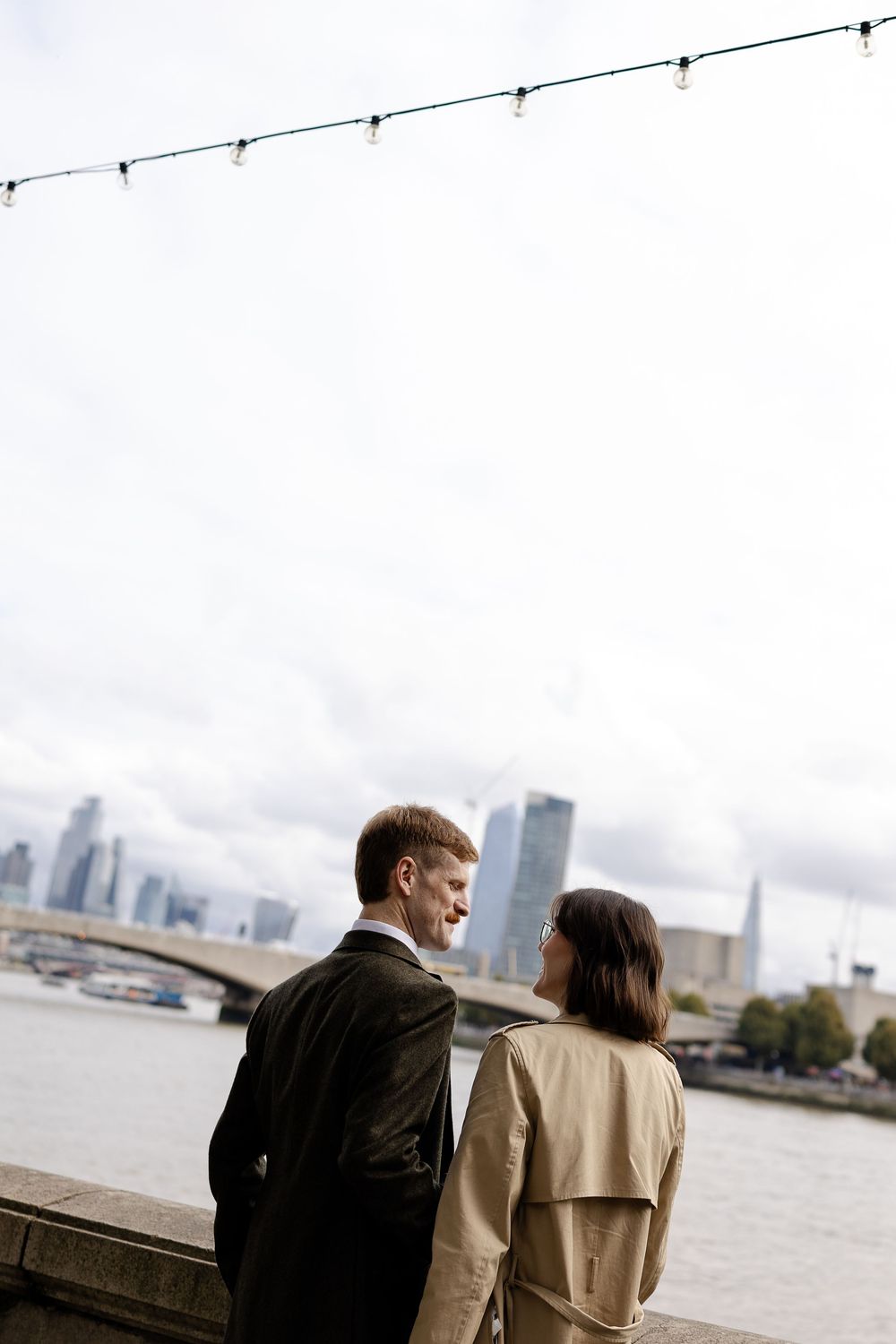 A couple in coats stands together looking out over a waterfront with city buildings visible in the distance.