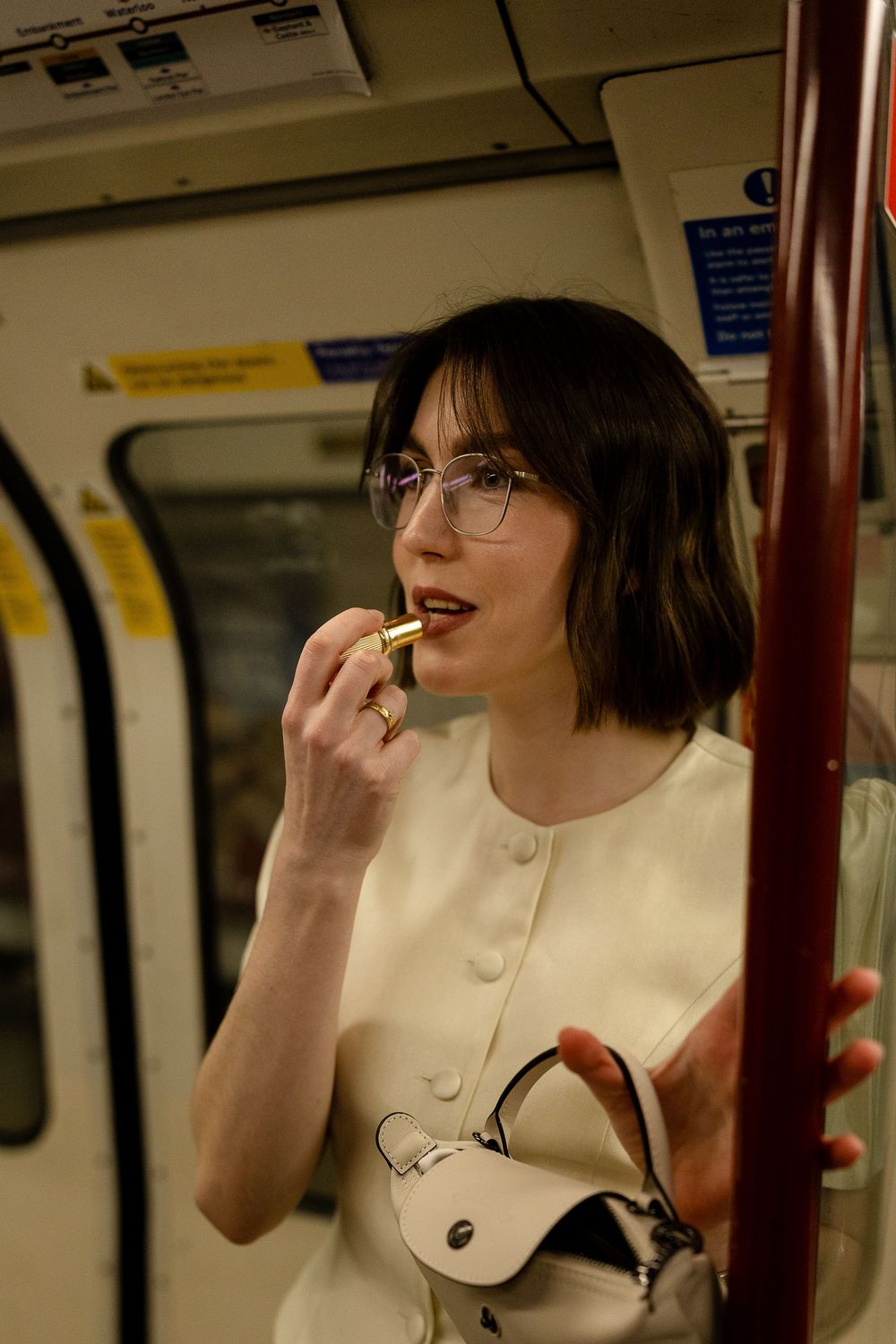 A person in a white top sits on public transit and applies lipstick.