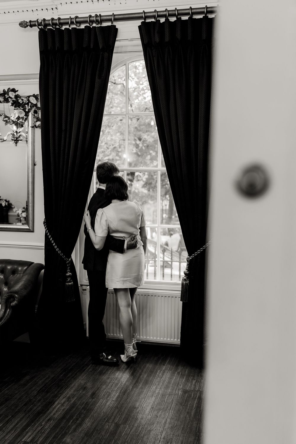 Black and white interior scene showing a figure in light clothing against dark curtains and hardwood floors.