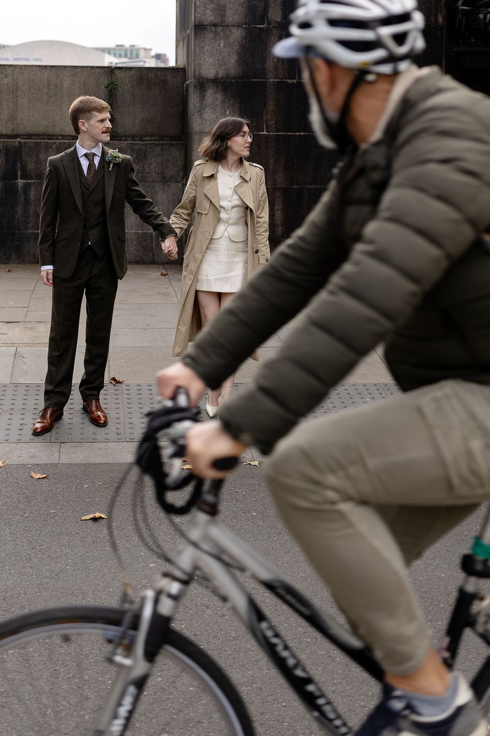 A cyclist rides past two people in formal attire standing on a city sidewalk.