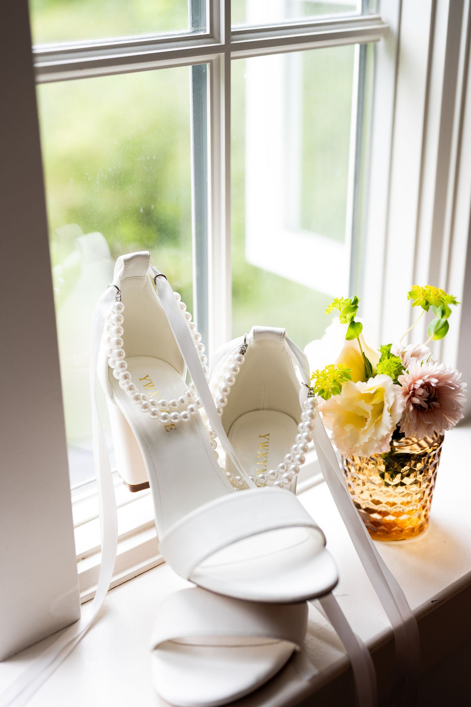 White pearl-strap bridal heels displayed on a windowsill beside a small amber vase with fresh flowers.