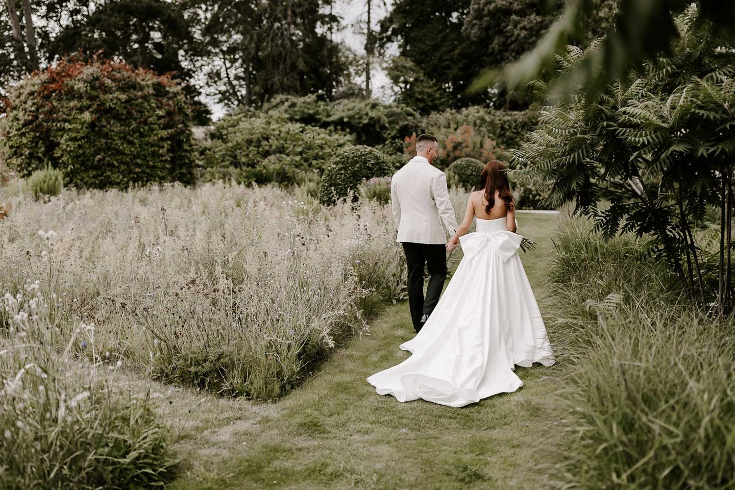Couple in wedding attire embracing in a rustic field of tall grass and wildflowers during golden hour.