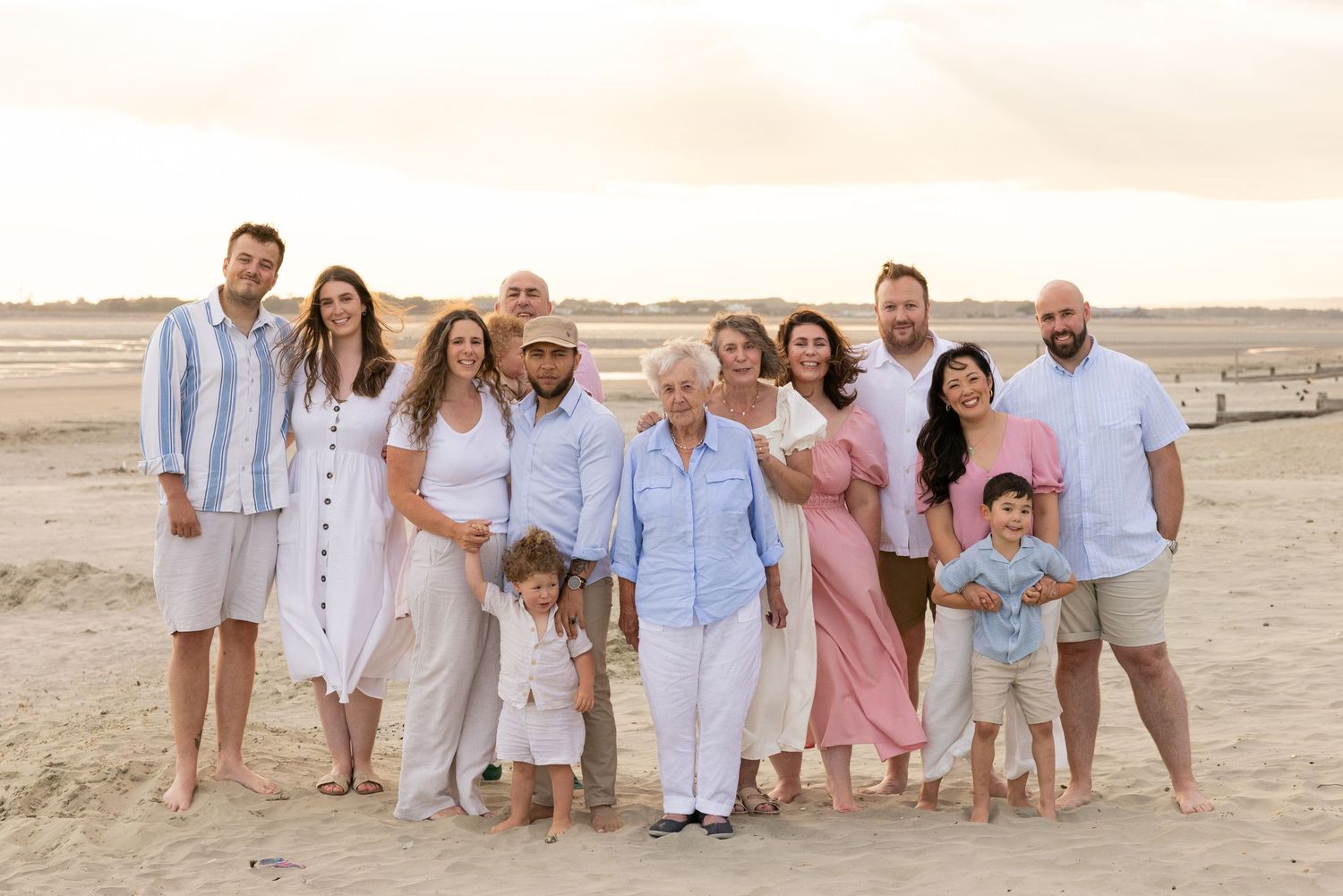 Family group wearing coordinated white and pastel outfits poses together on sandy beach at sunset.
