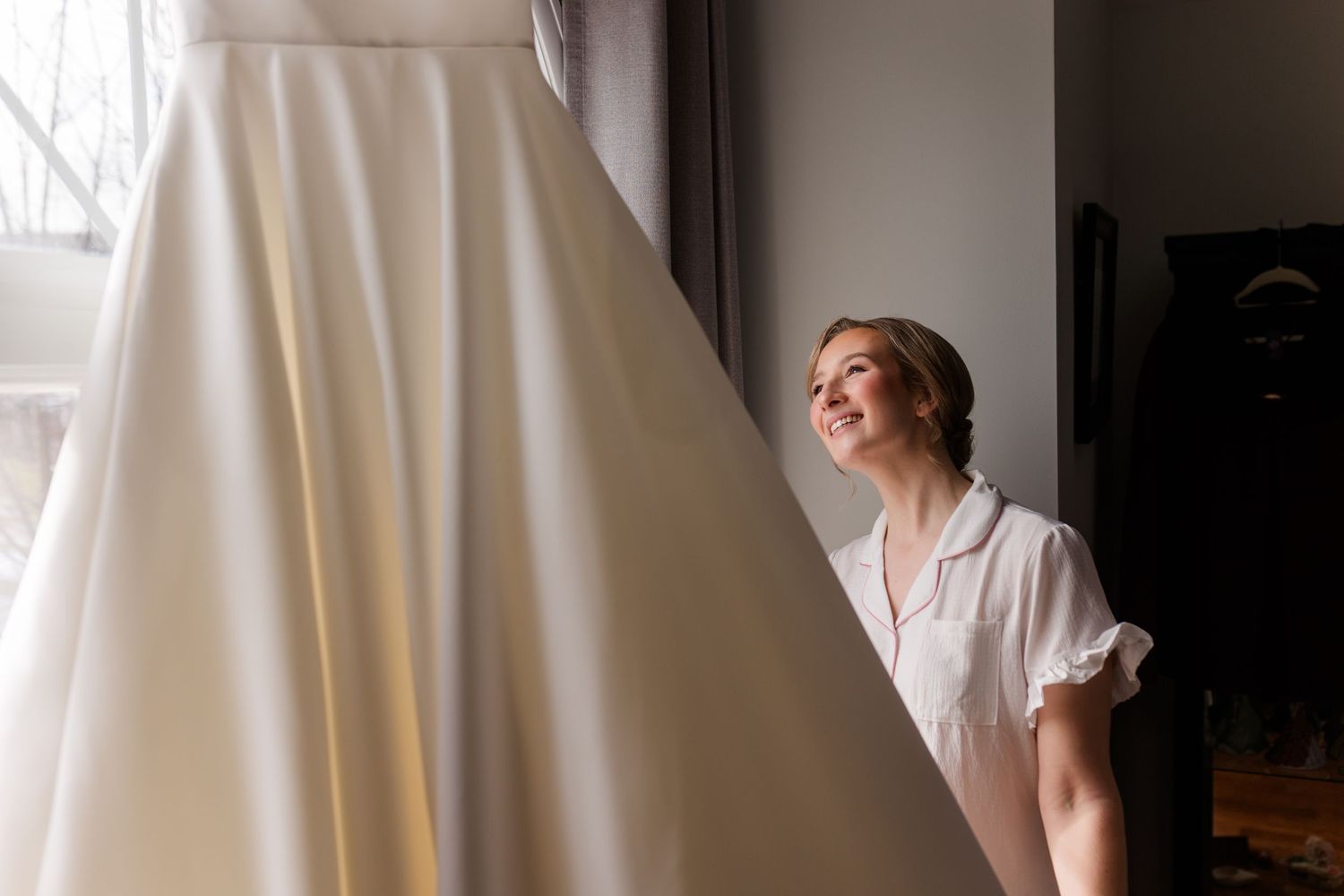 A bride in a white wedding dress stands by a window with soft natural light streaming in.