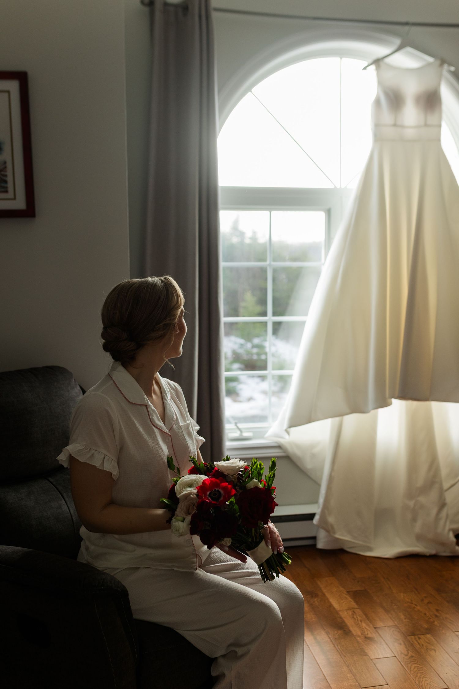A figure in a white dress sits by a window holding red flowers in a sunlit room with hardwood floors.