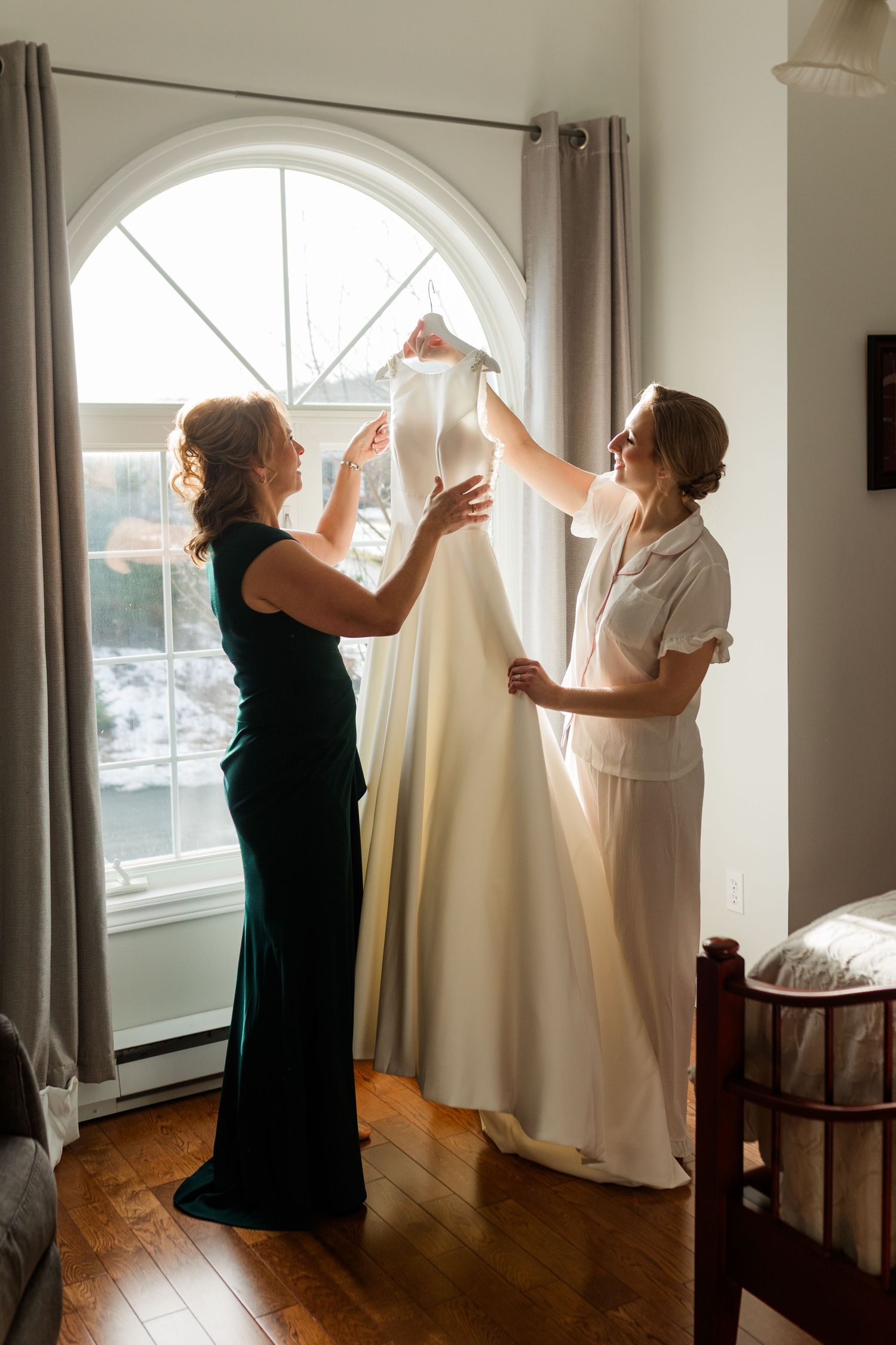 Two people hang white curtains on tall arched windows in a bright sunlit room with hardwood floors.