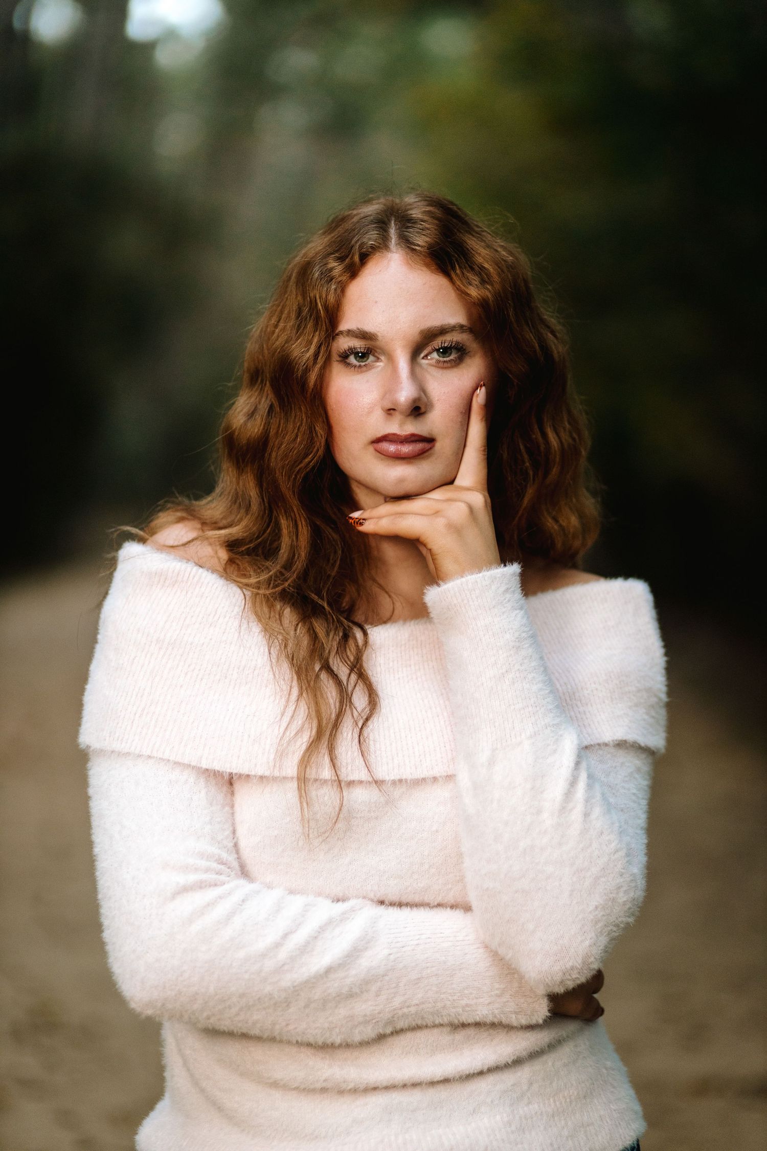 A person wearing a cozy white sweater poses outdoors with red lipstick and wavy hair in a natural setting.