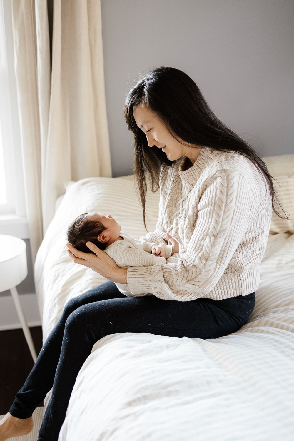 A mom smiles at her new baby while sitting on a bed at her San Jose home