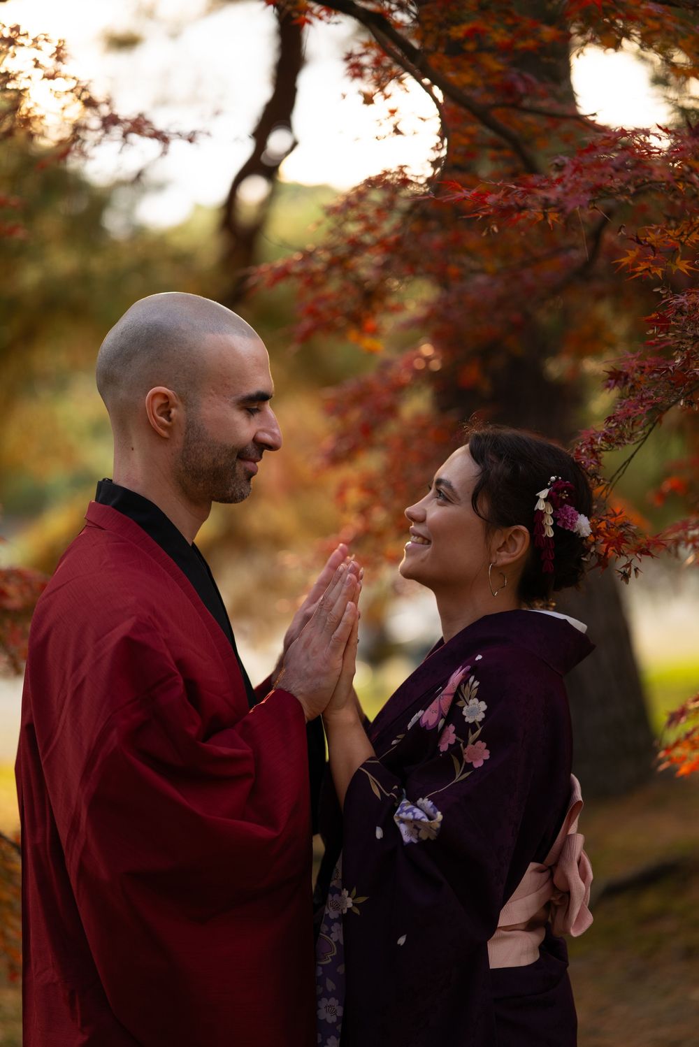 Smiling couple in traditional Japanese kimono pressing palms together in a playful pose under autumn maple trees.