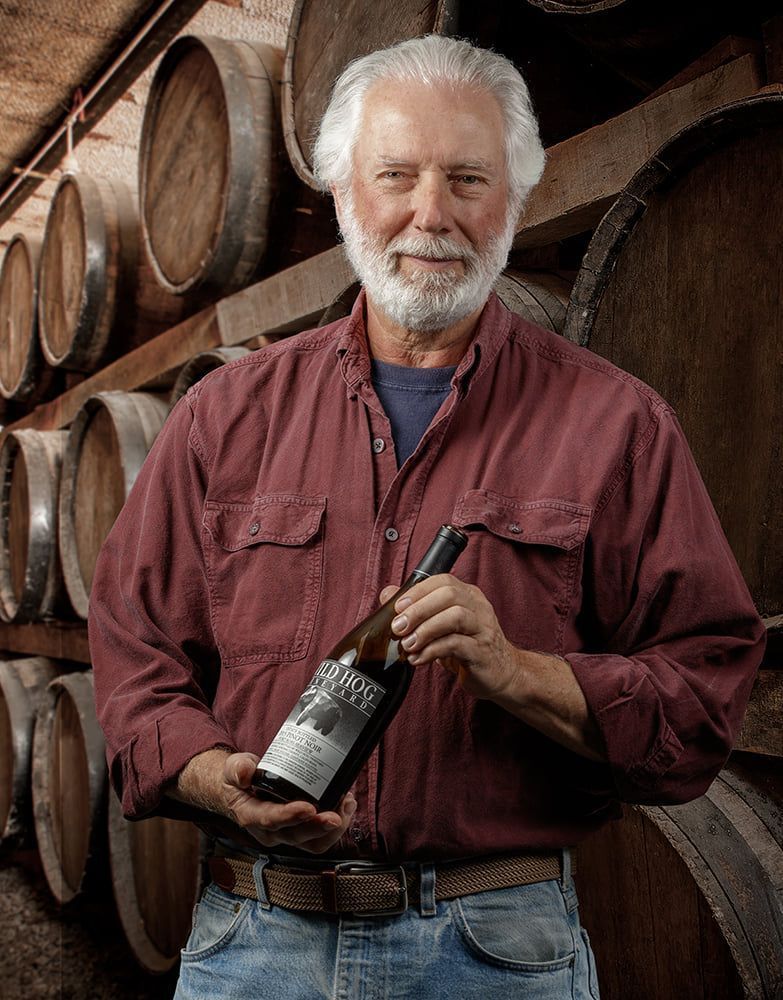 Winemaker displays bottle in cellar with oak barrels in background.