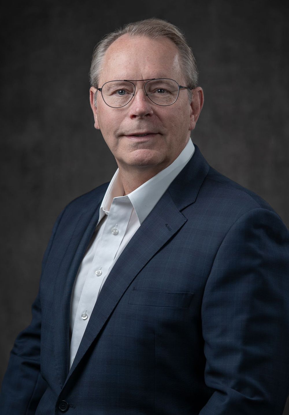 Professional headshot portrait of someone in a navy suit jacket and white dress shirt wearing glasses against dark background.