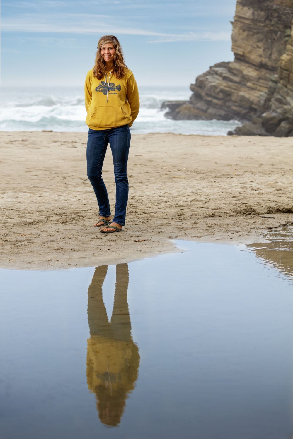 A person in a yellow hoodie walks along a coastal beach with dramatic cliffs and their reflection in a tidal pool.