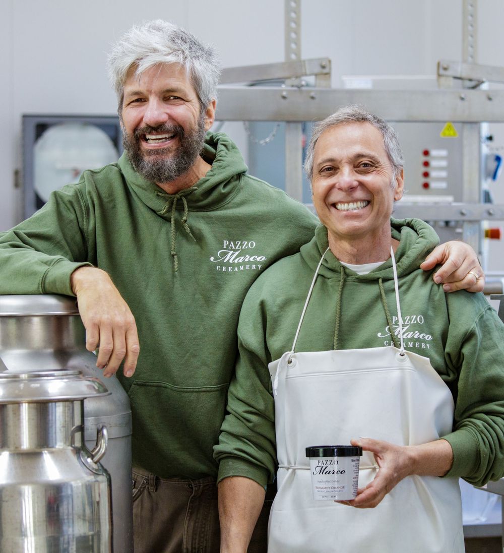 Two people wearing green hoodies smile while working in a commercial kitchen setting.