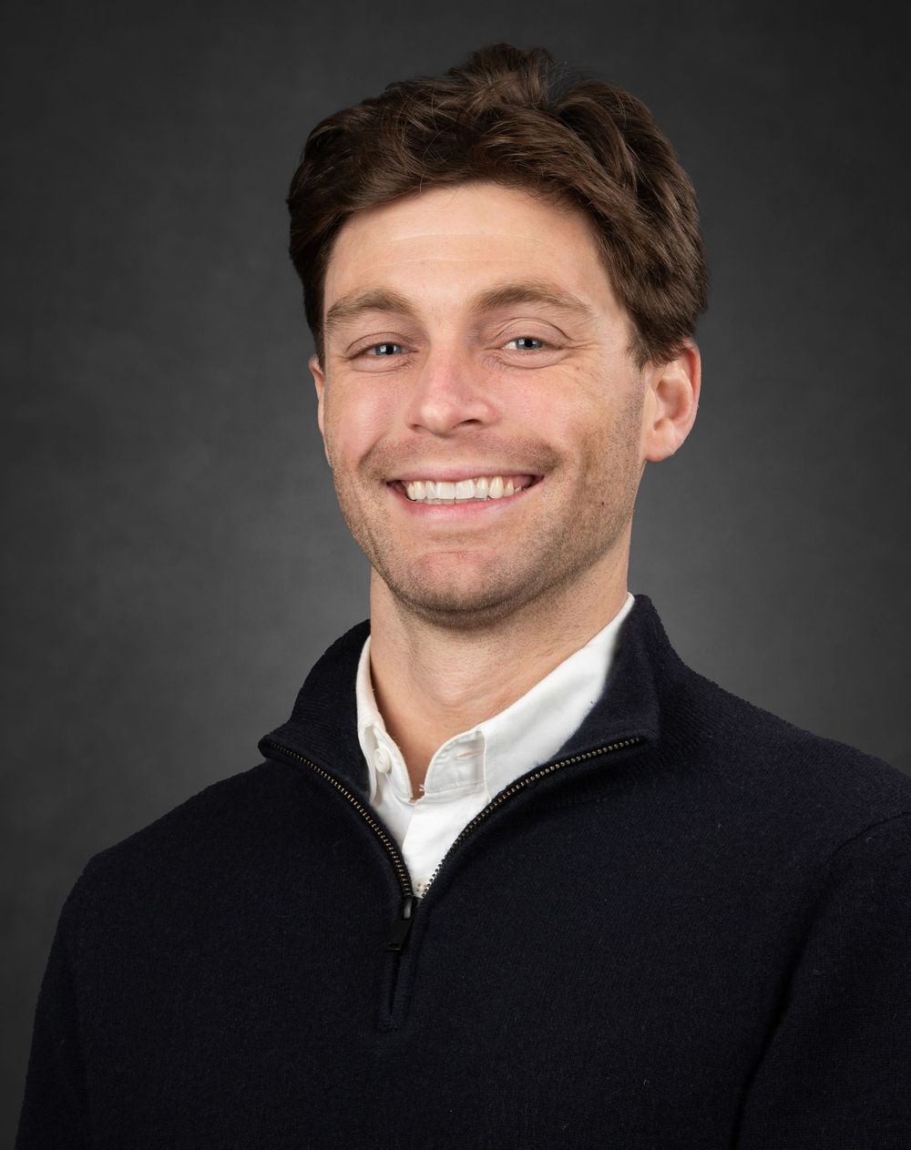 Professional headshot portrait against dark background showing warm smile and black coat.