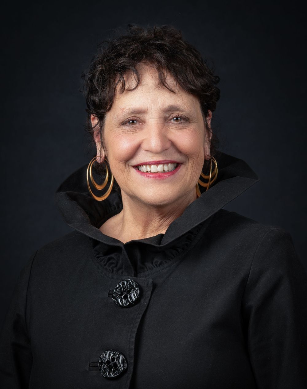 Professional portrait against dark background featuring gold hoop earrings and black jacket.