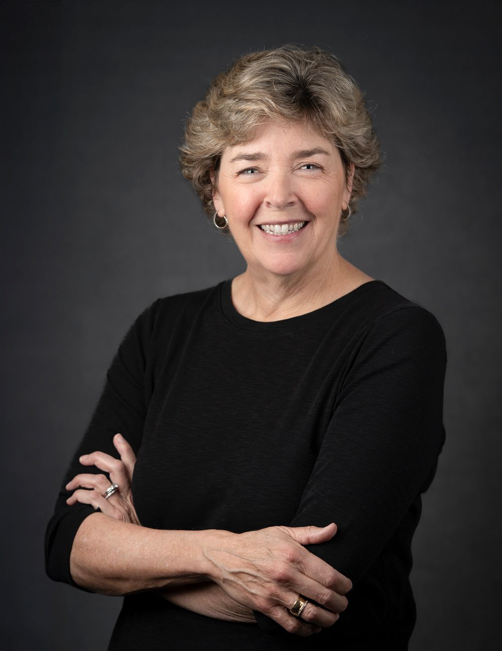 Professional headshot portrait of a person smiling warmly against a dark background wearing a black sweater.