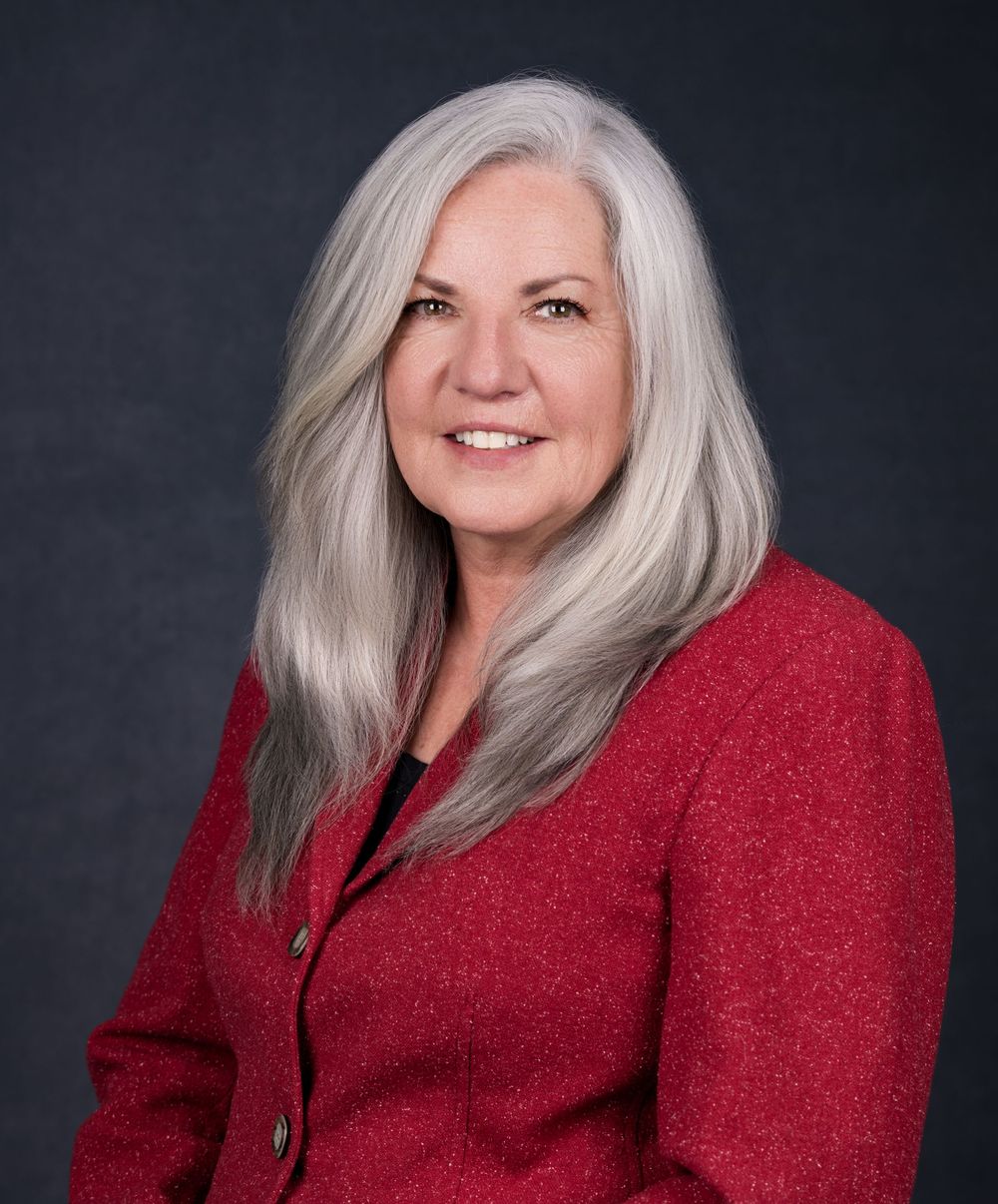 Professional woman with long gray hair wearing a red blazer, smiling confidently against a dark background.
