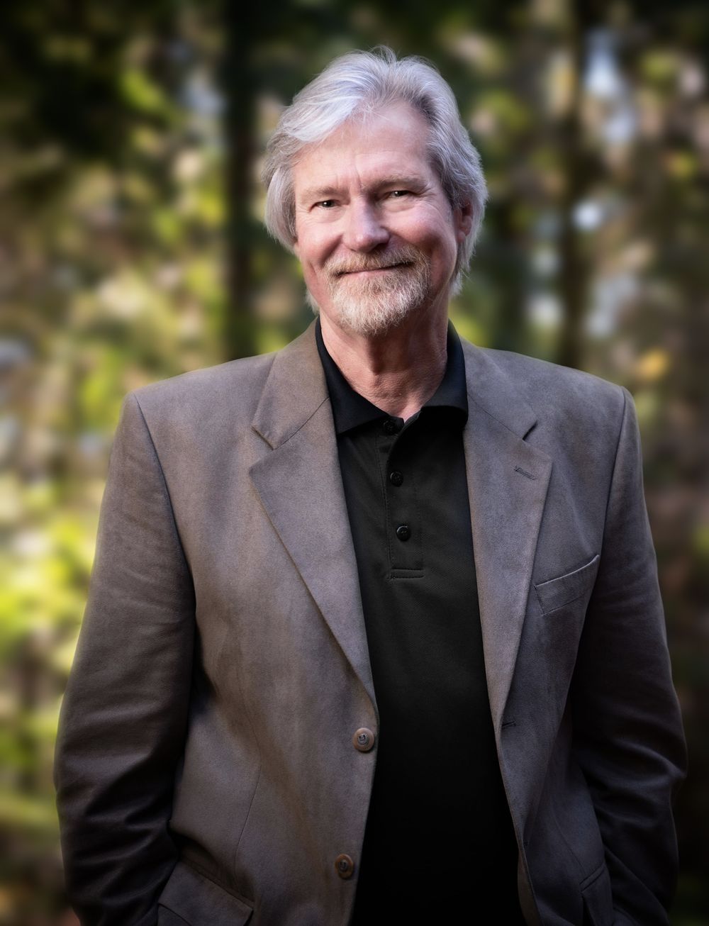Portrait of distinguished man in business attire smiling warmly against natural outdoor setting.