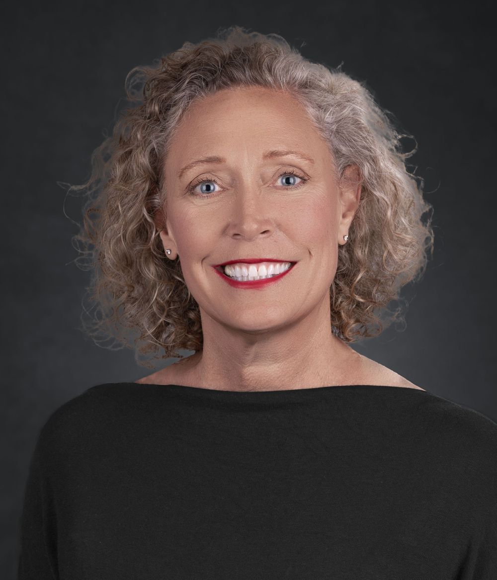 Professional headshot portrait of a person with curly gray hair wearing a black top against a dark background.