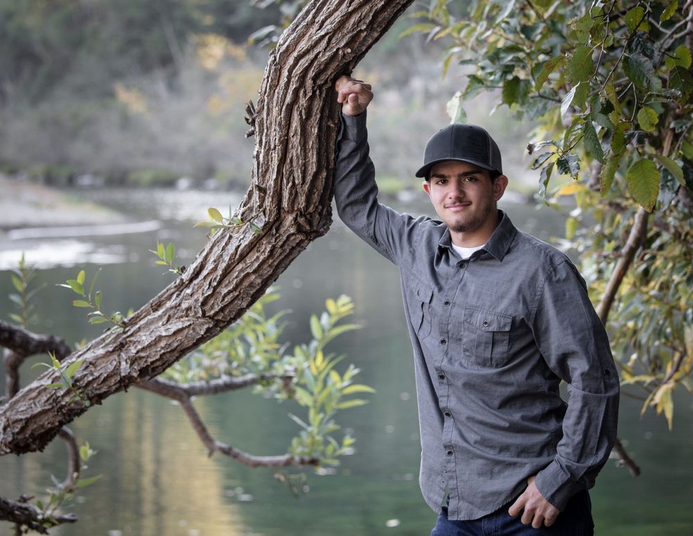 A person wearing a dark jacket sits peacefully by a lake surrounded by overhanging tree branches and calm water.