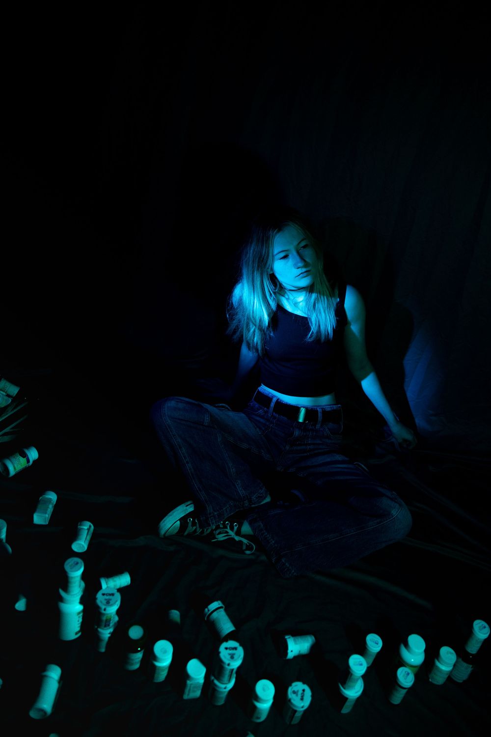 A figure sits cross-legged in dark clothing surrounded by bottles under ethereal blue lighting.