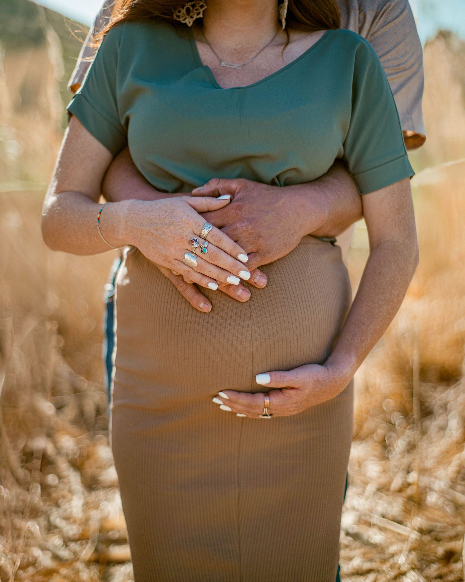 Close-up of hands cradling pregnant belly in brown dress surrounded by wheat stalks.