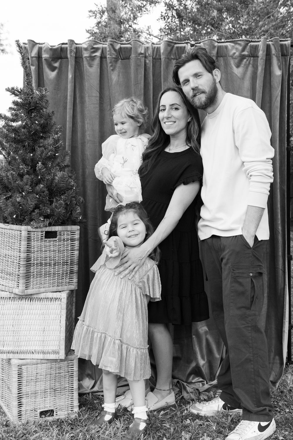 Black and white family portrait of a bearded man, a smiling woman holding a toddler, and a little girl beside a Christmas tree.