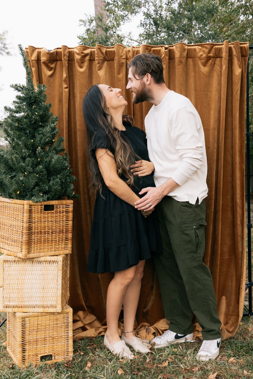 Expecting couple embraces and smiles at each other in front of a golden velvet backdrop beside a small Christmas tree in a basket.