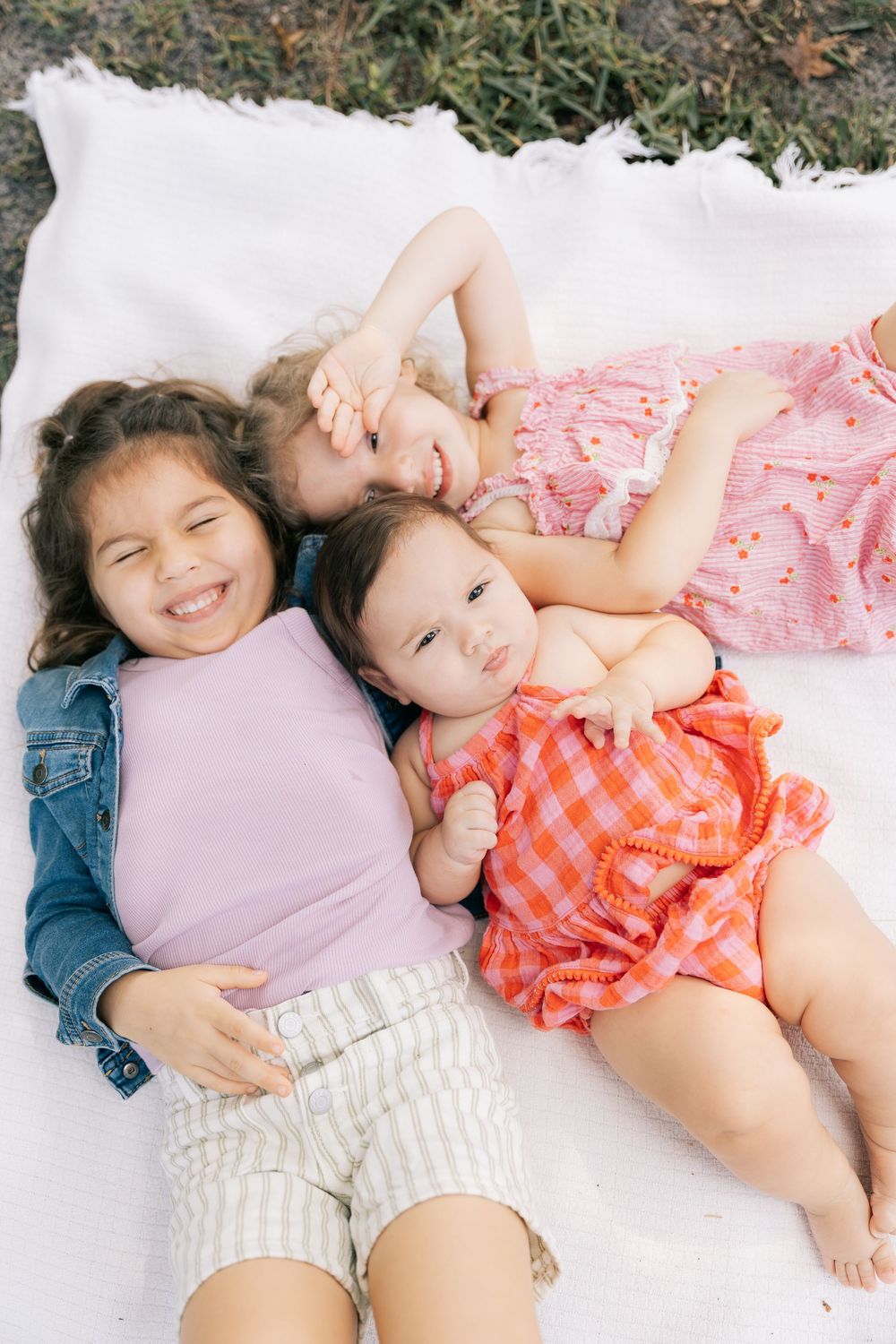 Three smiling young sisters lie together on a white blanket in the grass, seen from above in a cheerful family portrait.