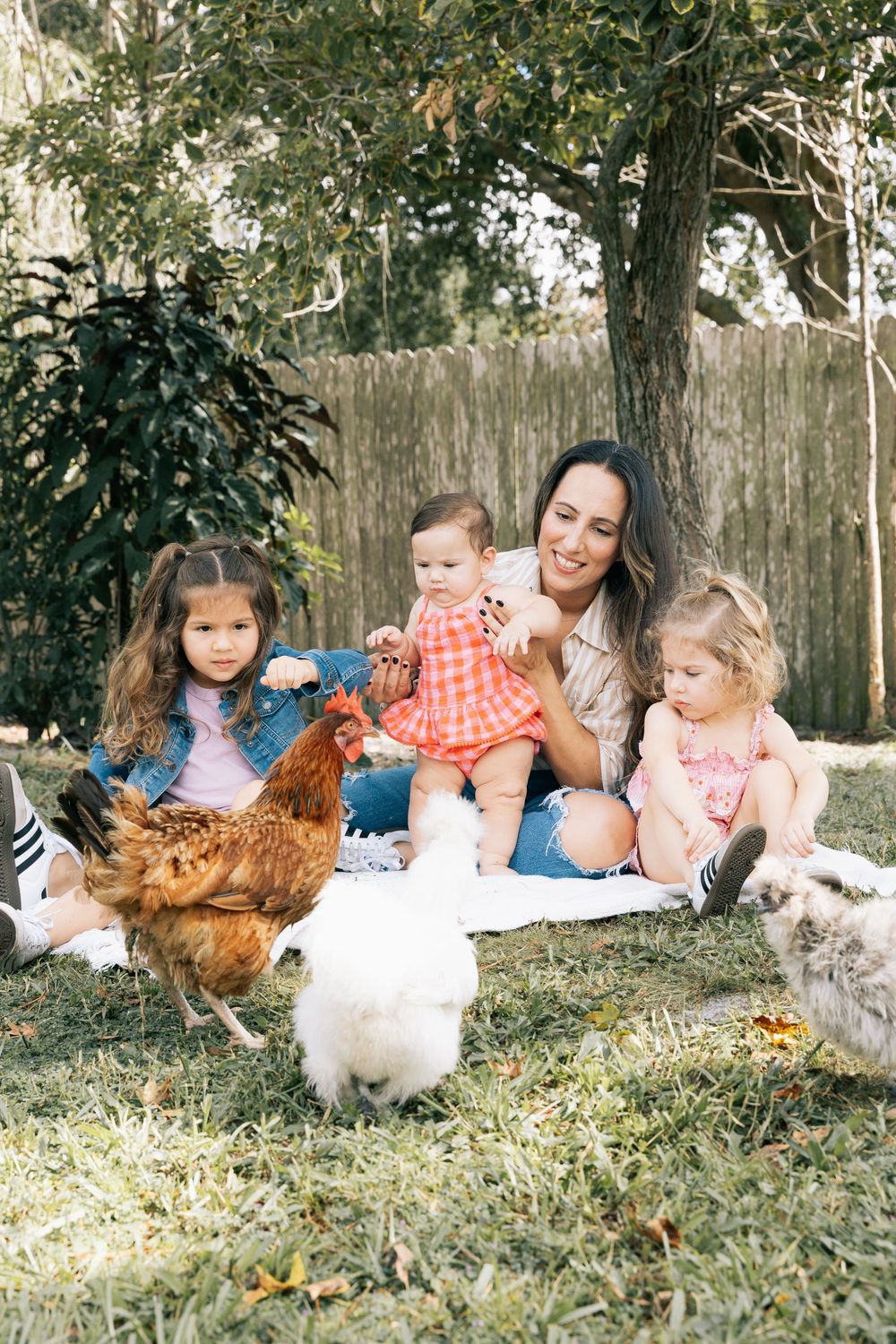 Mother and three young daughters sit on a blanket in a backyard watching chickens roam the grass.