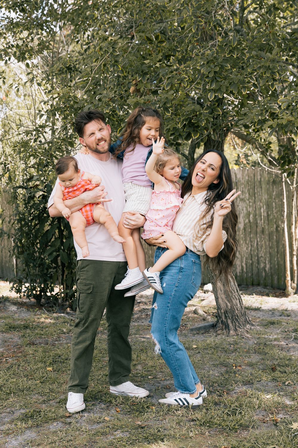 Smiling family of five poses outdoors, with a father holding a baby and a daughter while the mother laughs and flashes a peace sign.