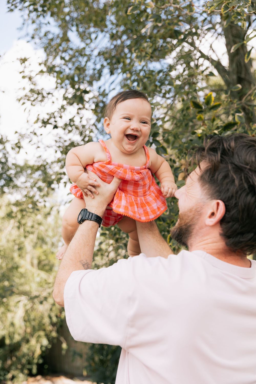Smiling father lifts a laughing baby girl in a red checkered dress high into the air outdoors beneath leafy trees.