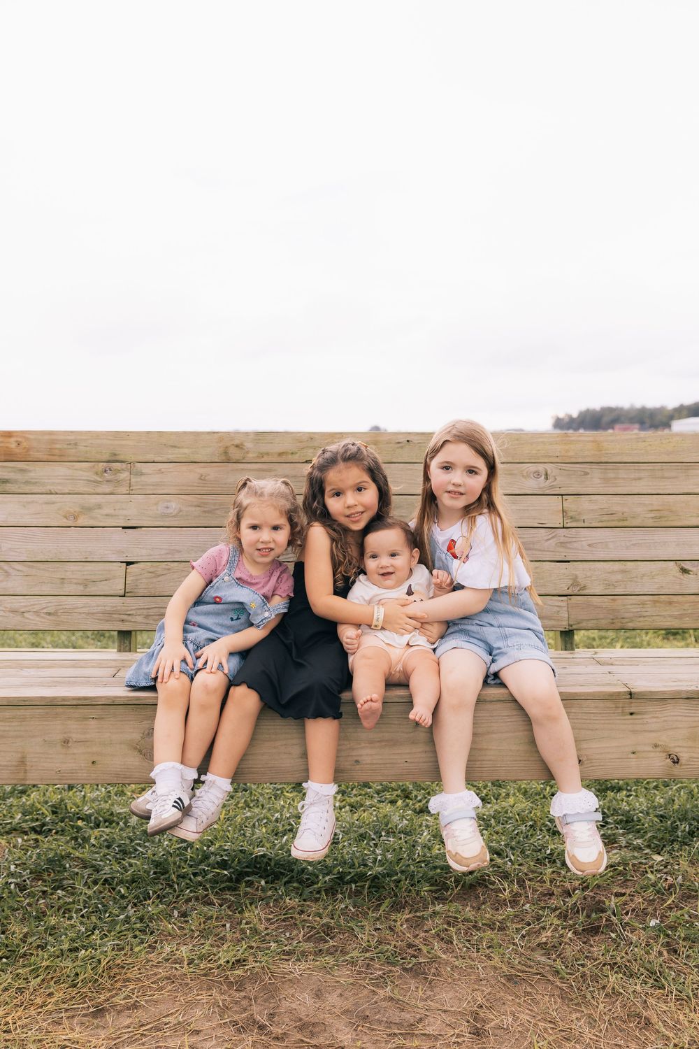 Four young sisters sit close together on a wooden bench outdoors, smiling, with a baby held in the middle.