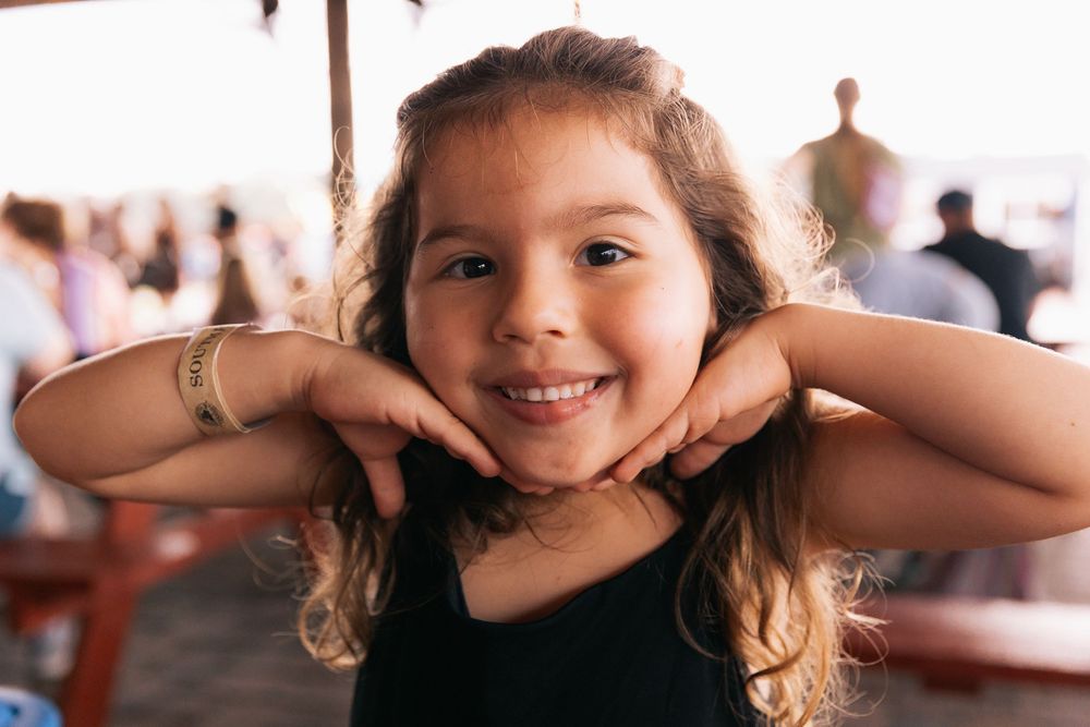 Smiling little girl rests her chin on her hands at an outdoor gathering, wearing a black top and gold bracelets.