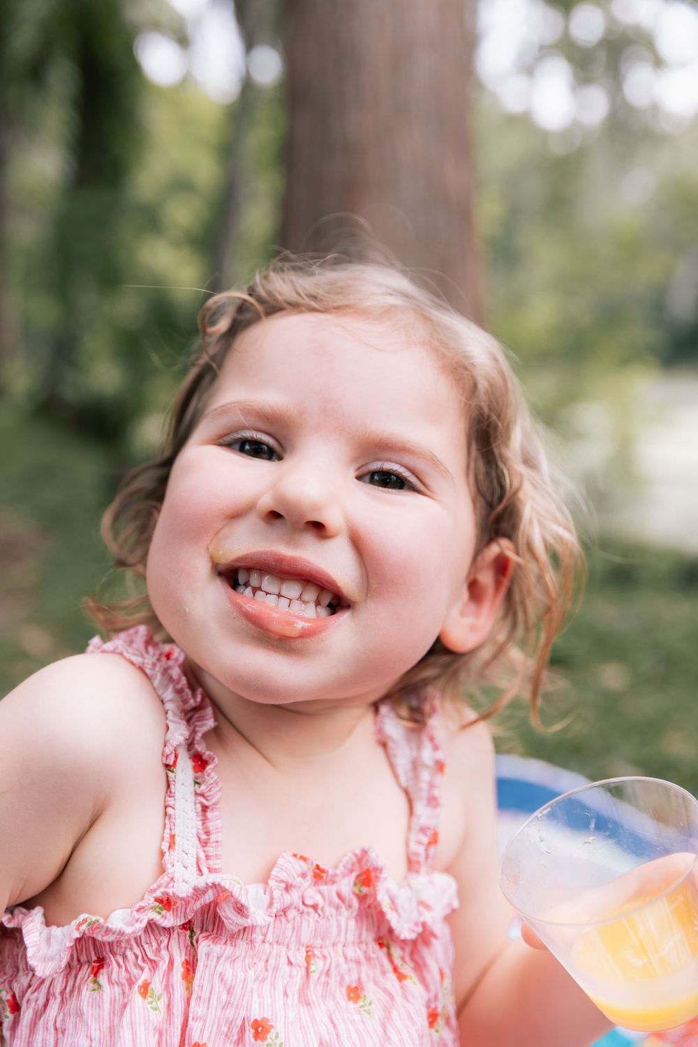 Giggling toddler girl in a pink floral dress sticks out her tongue outdoors among trees while holding a cup of juice.
