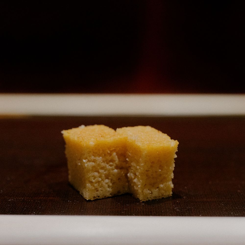 A small piece of yellow cornbread with a bite taken out sits on a dark surface.