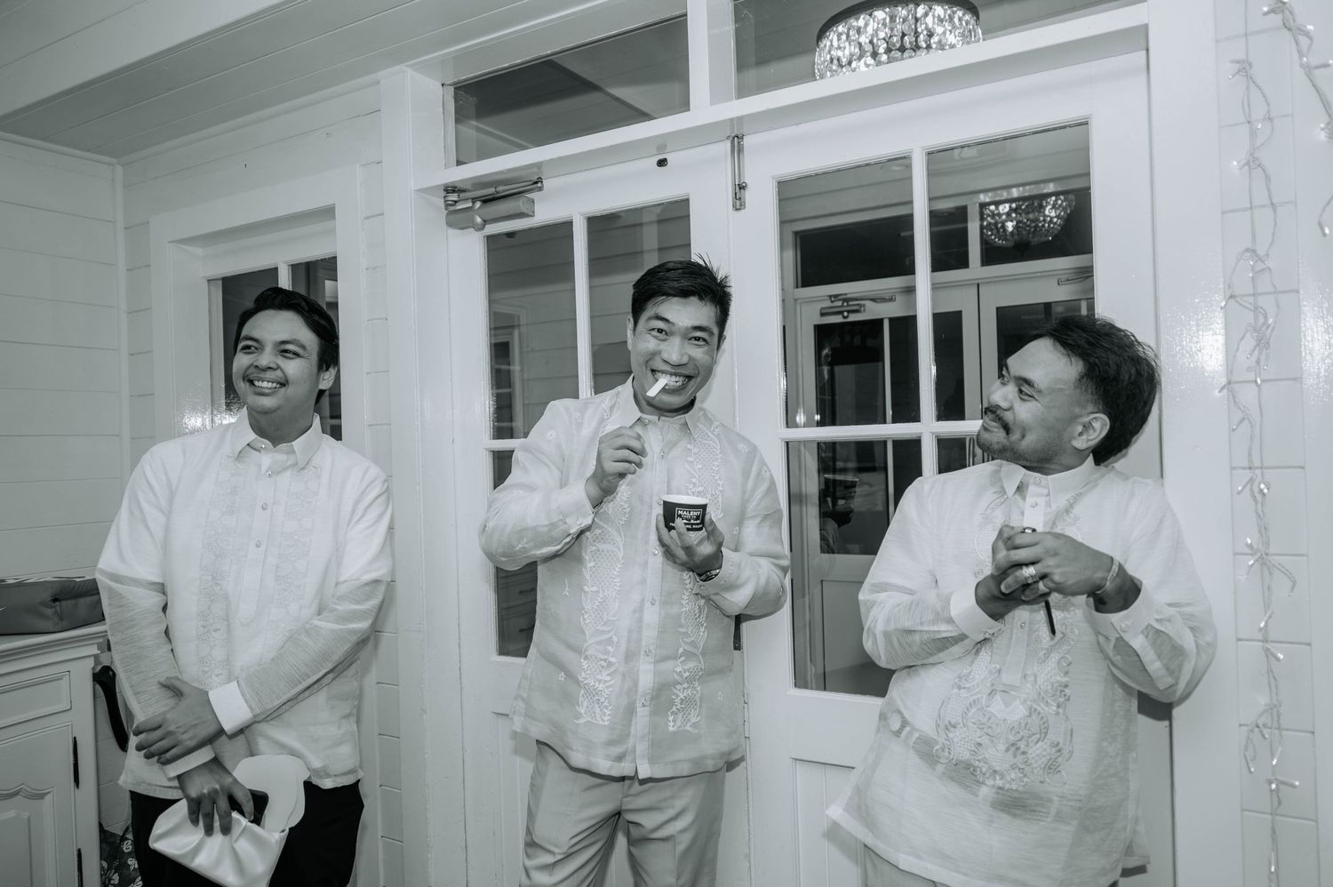 Black and white candid photo of three groomsmen sharing laughs outside a white building.