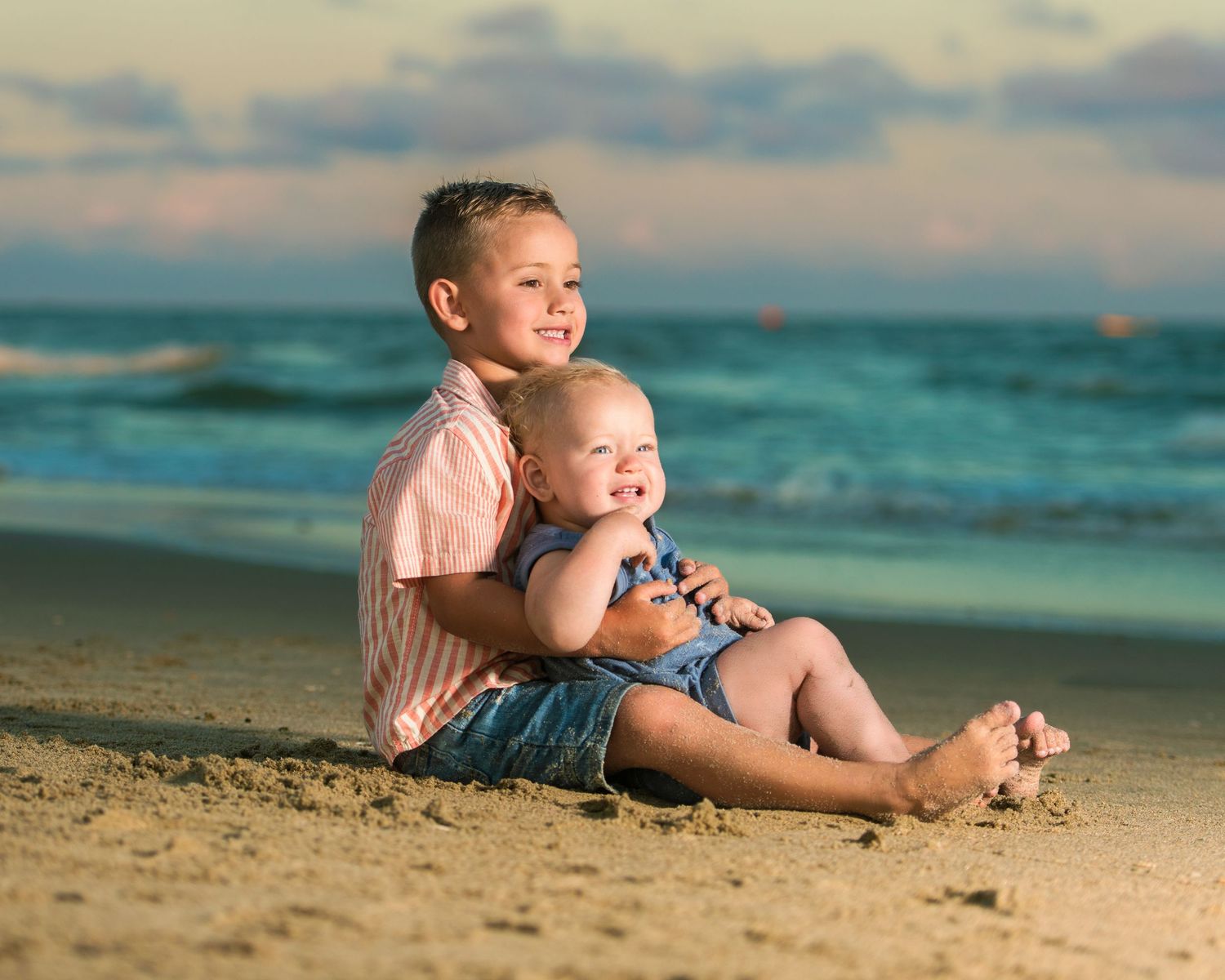 Two children sitting together on a sandy beach at sunset with turquoise ocean waves in the background.