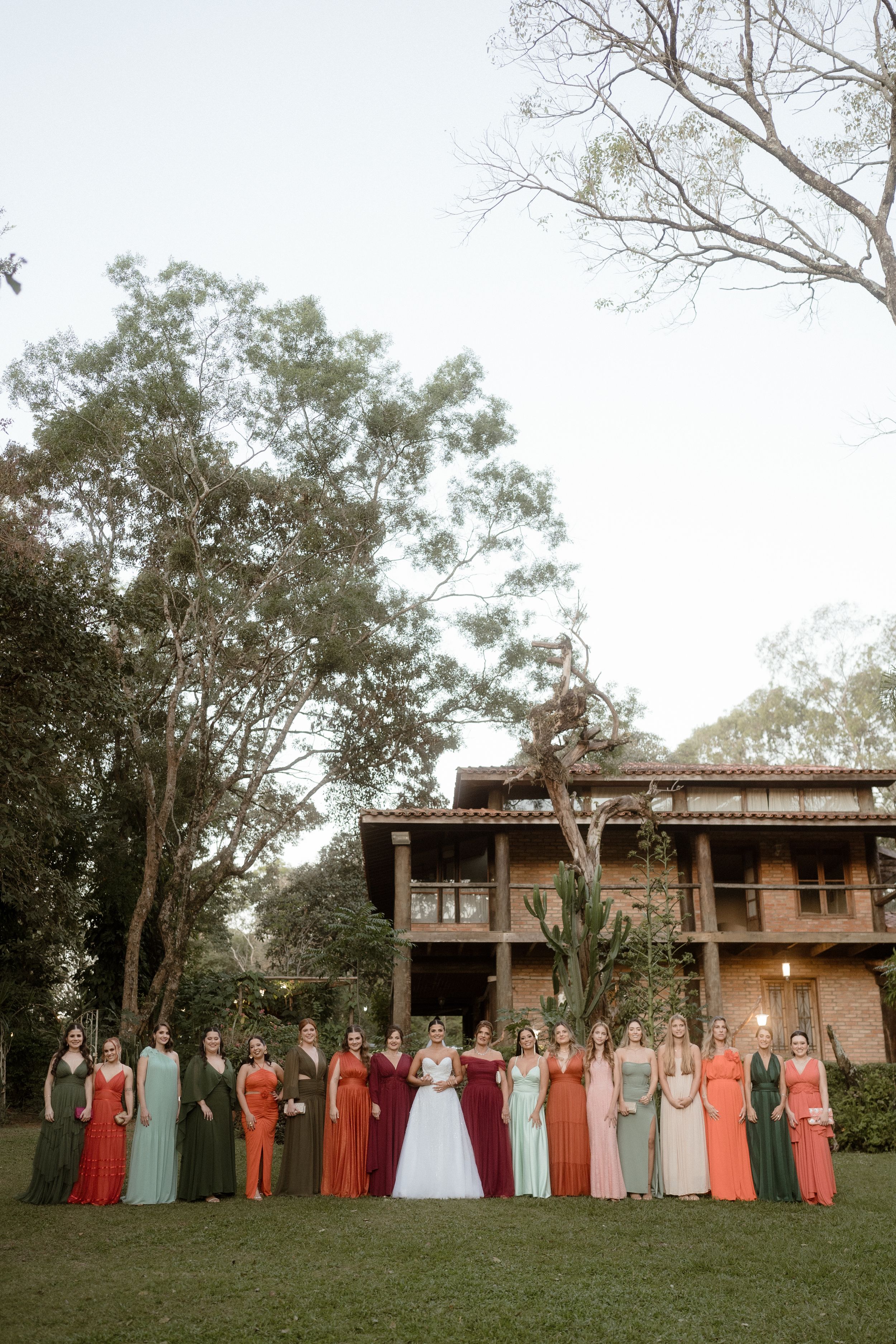 Wedding party in colorful dresses poses in front of a wooden building surrounded by trees.