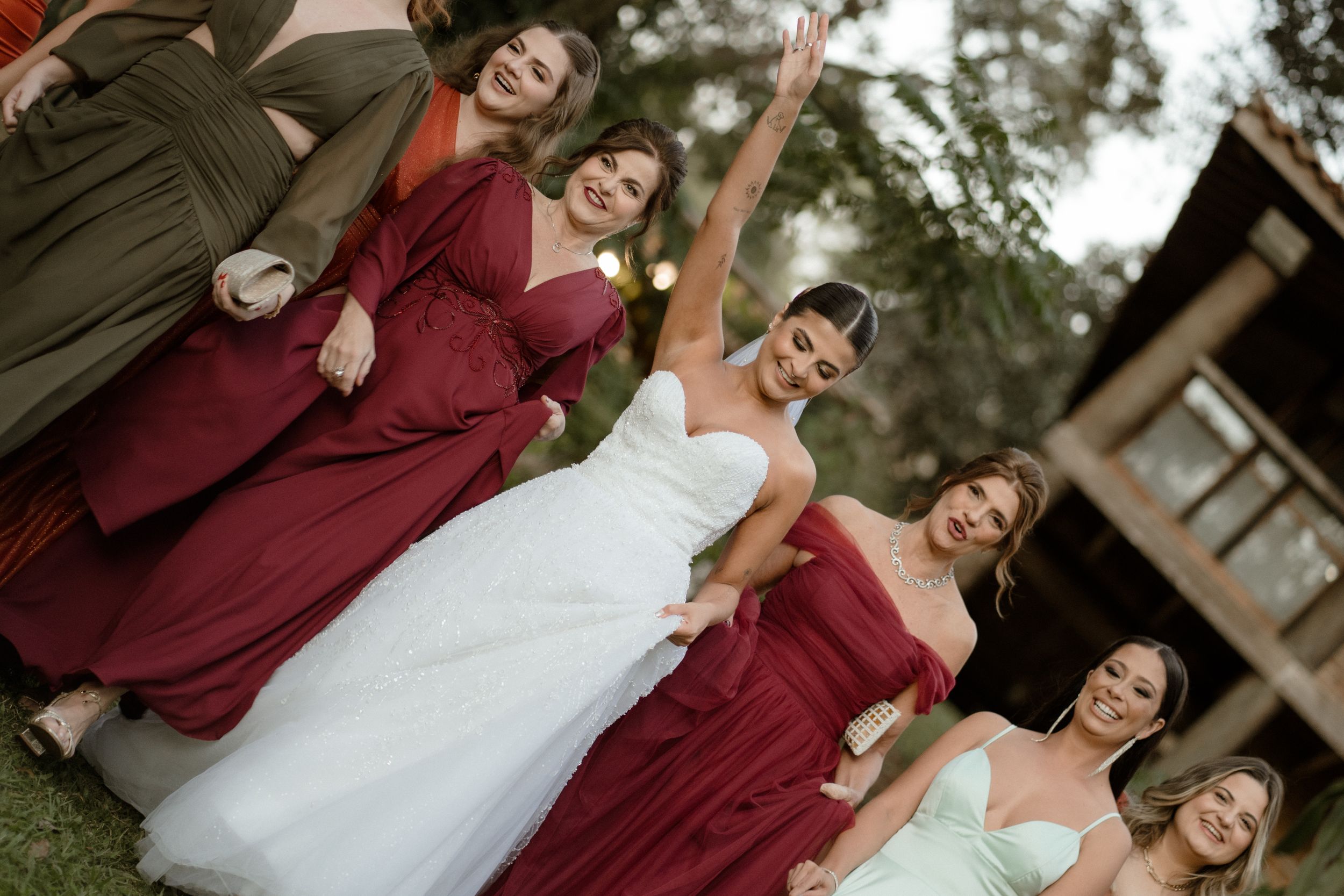 Wedding party poses joyfully in burgundy dresses and white gown against rustic backdrop.