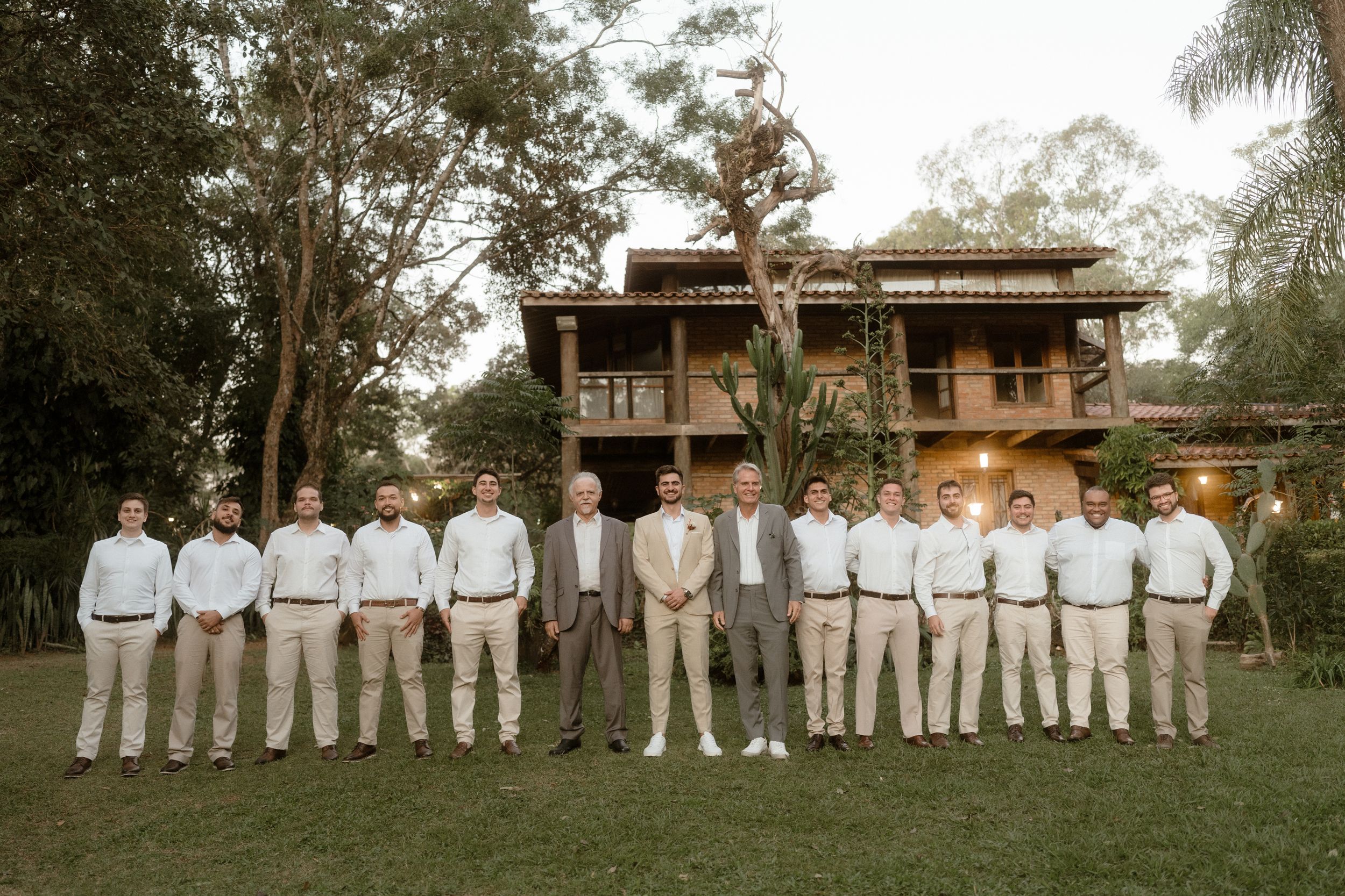 Groomsmen in light colored suits line up on lawn in front of wooden building.